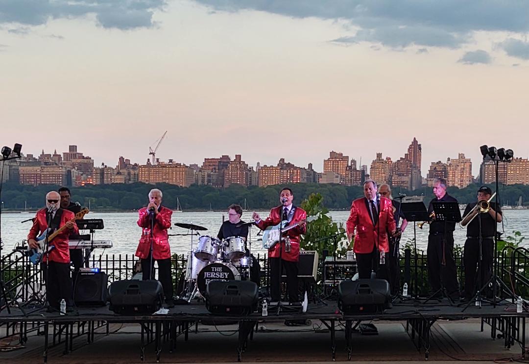 The Jersey Four on an outdoor stage by the Hudson, with the New York skyline behind them